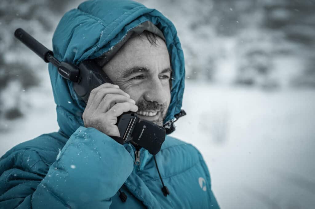 A man in a snowy hooded jacket holding a handheld radio to his ear, antenna extended above his hood